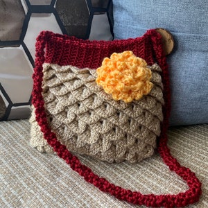 May include: A crocheted handbag with a red strap and a yellow flower accent. The bag is made with a textured, scale-like pattern.