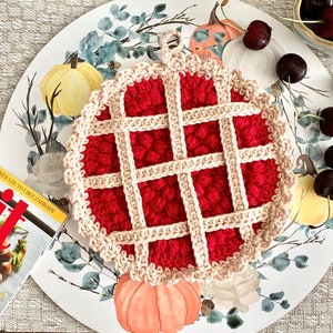 May include: A crocheted red and cream pie-shaped hot pad with a loop handle. The hot pad is on a white plate with a floral design and surrounded by cherries.