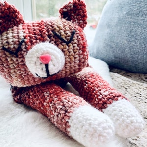 May include: A crocheted cat toy with a pink nose, black eyes, and white paws. The cat is lying on a white fluffy surface.