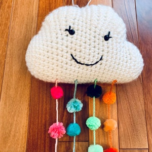 May include: A white knitted cloud decoration with a smiling face and hanging pom-poms. The pom-poms are in various colours, including pink, teal, green, orange, and black. The cloud has a loop for hanging, and the overall effect is a rainbow.