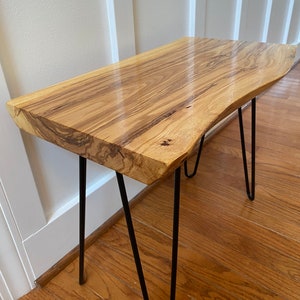 May include: A small, live edge wooden table with a natural wood grain finish. The table has black hairpin legs. The table is approximately 30 inches long and 12 inches wide.