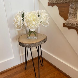 May include: A small wooden table with a natural bark edge and black hairpin legs. A clear glass vase with white peonies sits on the table.