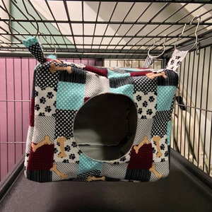 Puede incluir: Una cama para mascotas de tela patchwork con estampado de huesos y una gran abertura para la entrada. La cama está colgada de una jaula con ganchos de metal.