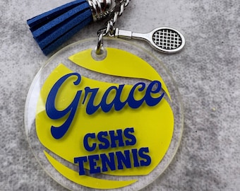 May include: A yellow tennis ball keychain with a blue tassel, a silver tennis racket charm, and the text "Grace CSHS Tennis".