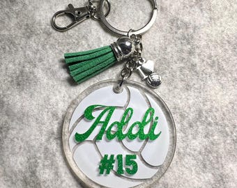 May include: A circular clear acrylic keychain featuring a white volleyball graphic with the green glitter text Addi and #15. The keychain includes a silver metal keyring, a lobster clasp, a green suede tassel, and a small silver heart and volleyball charm.
