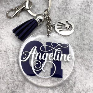 May include: A circular clear acrylic keychain featuring the dark blue letters OT and the white script name Angeline. It includes a dark blue tassel, a silver-toned metal keyring with a lobster clasp, and a round silver charm with a handprint cutout.