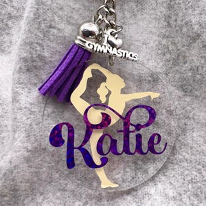 Può includere: Un portachiavi circolare in acrilico trasparente con la sagoma color crema di una persona che fa ginnastica. Il nome Katie è stampato in corsivo glitterato viola sulla figura. Include una nappa in camoscio viola e un ciondolo argentato.
