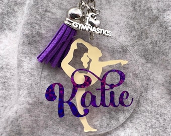 May include: A clear circular acrylic keychain featuring a cream-colored silhouette of a gymnast in a pose. The name Katie is printed in purple glitter script over the figure. A purple suede tassel and a silver charm reading I Heart Gymnastics.