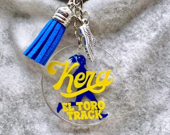 May include: A circular clear acrylic keychain featuring a blue silhouette of a runner, the yellow text "Kera" and "EL TORO TRACK", a bright blue tassel, and a silver-toned metal charm shaped like a running shoe attached to a metal ring.