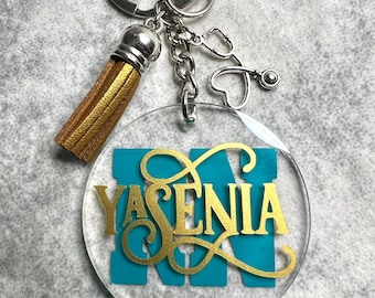 Può includere: Un portachiavi circolare in acrilico trasparente con il nome YASENIA in lettere dorate sovrapposte a una lettera N color verde acqua. Include una nappa dorata, un anello in metallo argentato e un ciondolo a forma di stetoscopio con cuore.