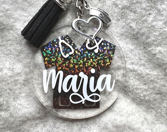 May include: A clear circular acrylic keychain featuring a black scrub top graphic with a rainbow glitter finish. The name Maria is printed in white script. It includes a black tassel, a silver heart charm, and a silver nurse cap charm.