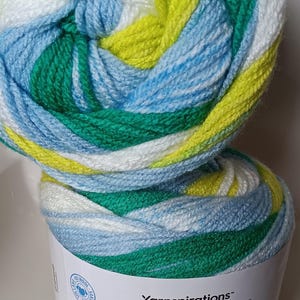 May include: A close-up of two Caron Cinnamon Swirl Cakes yarn skeins. The yarn is a blend of white, light blue, green, and yellow. The label reads "Caron Cinnamon Swirl Cakes" in colorful text. The yarn is 100% wool.