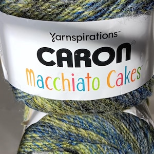 May include: A close-up of two skeins of Caron Macchiato Cakes yarn. The yarn is a blend of blue, green, and gray. The label reads "Caron Macchiato Cakes" in a colorful font. The yarn is tightly wound.