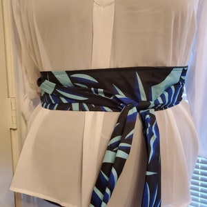 May include: A black and blue patterned fabric belt with a leaf design. The belt is tied around a white blouse.