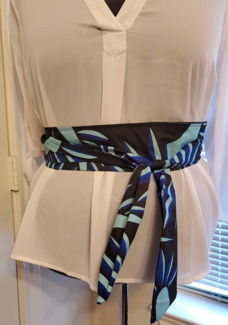 May include: A black and blue patterned fabric belt with a leaf design. The belt is tied around a white blouse.