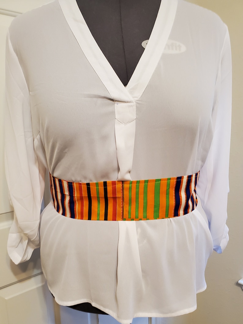 May include: A white long-sleeved blouse with a V-neck and a colorful patterned belt. The belt has stripes of orange, yellow, green, red, blue, and white.