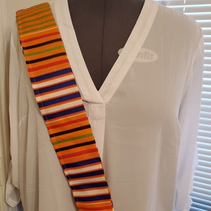 May include: A white blouse with a V-neckline and a colorful striped sash worn diagonally across the chest. The sash has orange, green, blue, white, and black stripes.