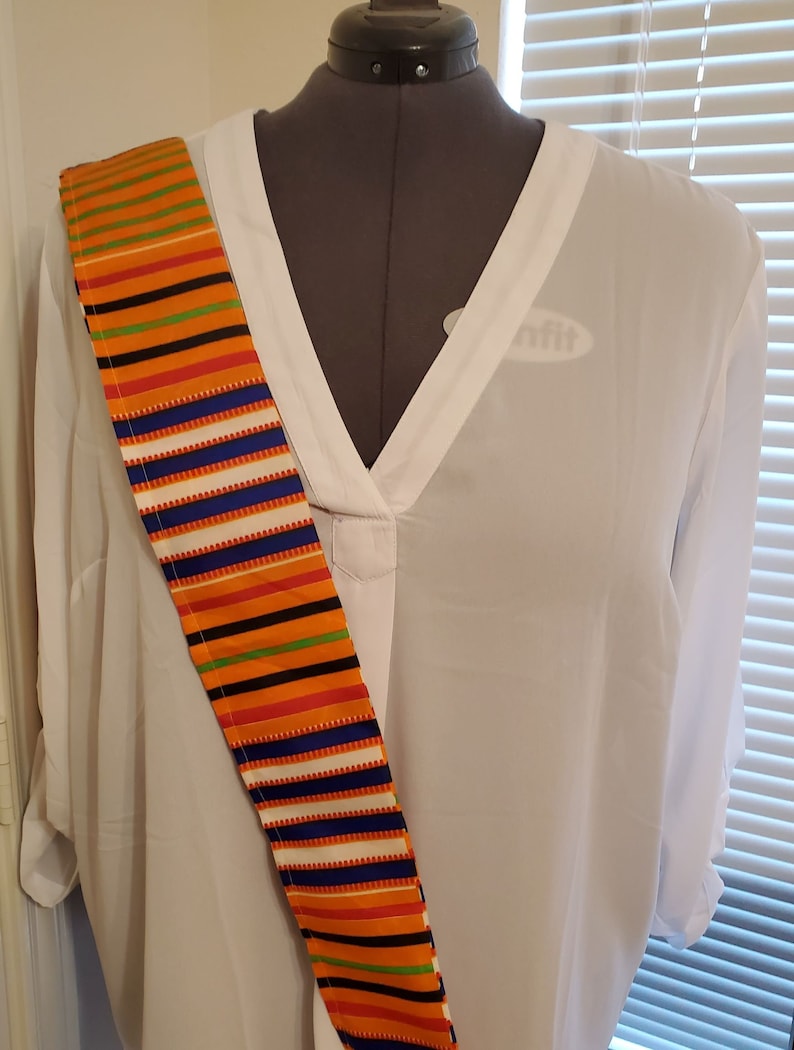 May include: A white blouse with a V-neckline and a colorful striped sash worn diagonally across the chest. The sash has orange, green, blue, white, and black stripes.