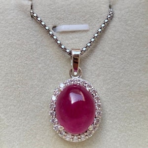 May include: A silver pendant necklace featuring an oval, deep red gemstone surrounded by small, clear stones. The pendant hangs from a delicate silver chain. The necklace is displayed against a neutral background.