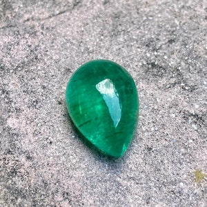 May include: A vibrant green emerald gemstone, cut into a teardrop shape, with a polished, reflective surface. The gemstone is set against a rough, grey background.