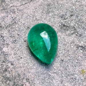 May include: A vibrant green emerald gemstone, cut into a teardrop shape, with a smooth, polished surface. The stone is set against a rough, grey background.
