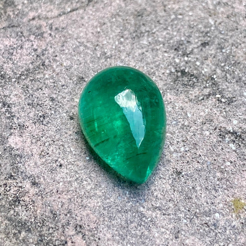 May include: A vibrant green emerald gemstone, cut into a teardrop shape, with a smooth, polished surface. The stone is set against a rough, grey background.