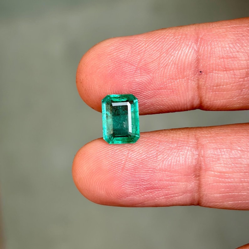 May include: A vibrant, rectangular emerald gemstone, held between fingers. The gem has a deep green hue and a polished surface, reflecting light. The emerald's shape is a classic rectangle, showcasing its clarity and color.