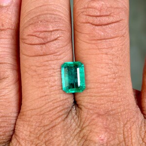 May include: A rectangular-cut emerald gemstone with a vibrant green hue. The gemstone is held between fingers, showcasing its clarity and brilliance. The emerald's facets reflect light, highlighting its polished surface. This gemstone is a beautiful example of natural emeralds.