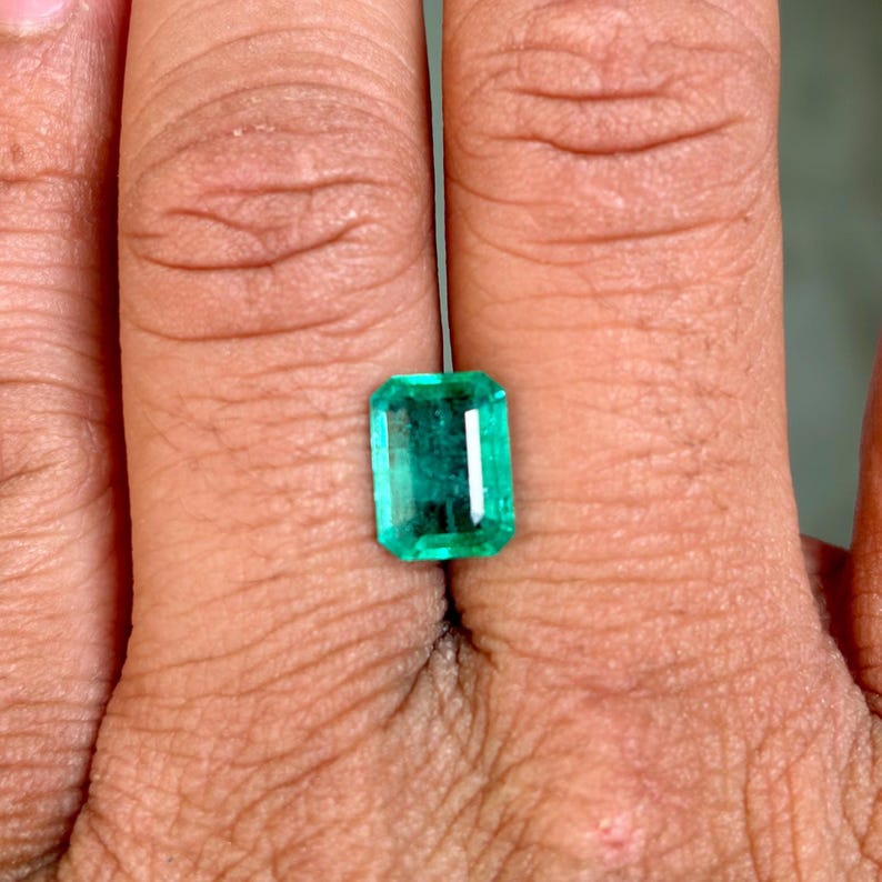 May include: A rectangular-cut emerald gemstone with a vibrant green hue. The gemstone is held between fingers, showcasing its clarity and brilliance. The emerald's facets reflect light, highlighting its polished surface. This gemstone is a beautiful example of natural emeralds.