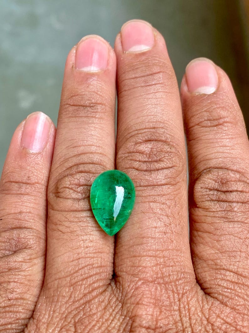 May include: A pear-shaped emerald gemstone with a deep green colour. The gemstone is set in a ring and is being held by a hand.