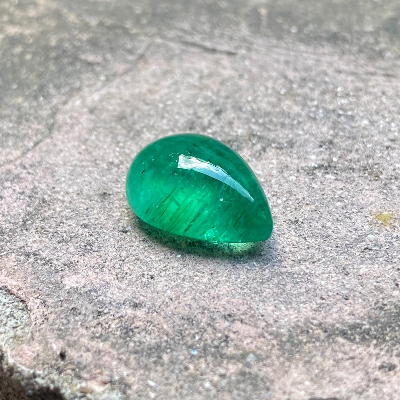 May include: A vibrant green emerald gemstone cut into a teardrop shape. The gemstone is faceted and has a high polish, reflecting light.