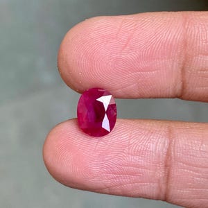 Puede incluir: Una gema de rubí facetada, de forma ovalada. El rubí es de color rojo intenso con facetas brillantes y reflectantes. La gema se sostiene entre dos dedos, mostrando su tamaño y brillo.