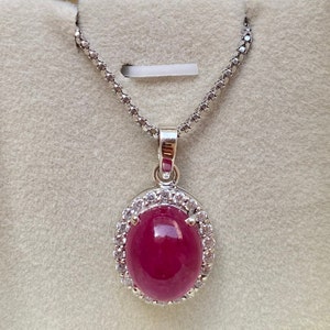 May include: A silver necklace with a pendant featuring a large, oval, ruby-colored gemstone. The gemstone is surrounded by a halo of small, clear gemstones. The chain is a delicate, linked design.