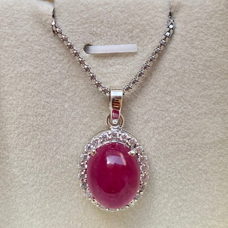 May include: A silver necklace with a pendant featuring a large, oval, ruby-colored gemstone. The gemstone is surrounded by a halo of small, clear gemstones. The chain is a delicate, linked design.