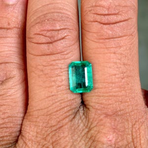 May include: A rectangular-cut emerald gemstone with a vibrant green hue. The gemstone is held in a hand, showcasing its clarity and color. The emerald's facets reflect light, highlighting its brilliance.