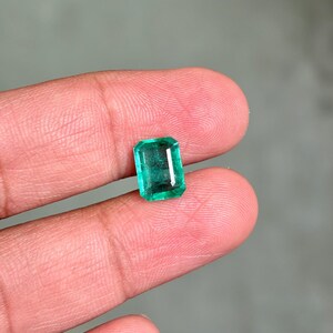 May include: A rectangular emerald gemstone with a vibrant green hue. The gem is held between two fingers, showcasing its clarity and cut. The emerald's facets reflect light, highlighting its brilliance. A close-up view emphasizes the gem's natural beauty.