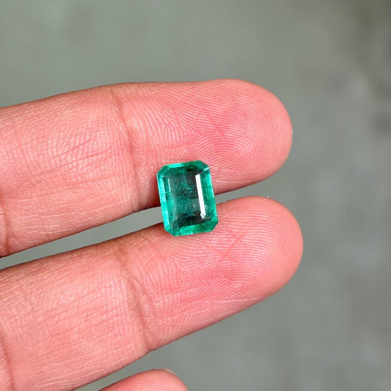 May include: A rectangular emerald gemstone with a vibrant green hue. The gem is held between two fingers, showcasing its clarity and cut. The emerald's facets reflect light, highlighting its brilliance. A close-up view emphasizes the gem's natural beauty.