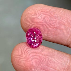 May include: A polished, oval-shaped, ruby gemstone with a carved face design. The gemstone is held between two fingers, showcasing its vibrant pink and red hues. The carving details a face with a smiling expression.