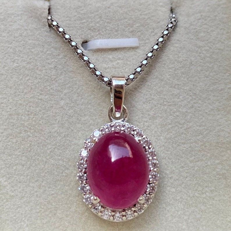 May include: A silver pendant necklace featuring an oval, deep red gemstone surrounded by small, clear stones. The pendant hangs from a delicate silver chain. The necklace is displayed against a neutral background.