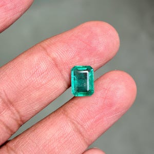 May include: A vibrant, rectangular emerald gemstone with a deep green hue. The gem is held between two fingers, showcasing its clarity and cut. The emerald's facets reflect light, highlighting its brilliance. This gemstone is a beautiful example of natural emeralds.