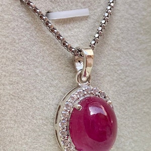 May include: A silver pendant necklace featuring a large, oval-shaped, deep red gemstone, surrounded by a halo of small, clear stones. The pendant hangs from a delicate, silver chain. The necklace is displayed against a light gray background.