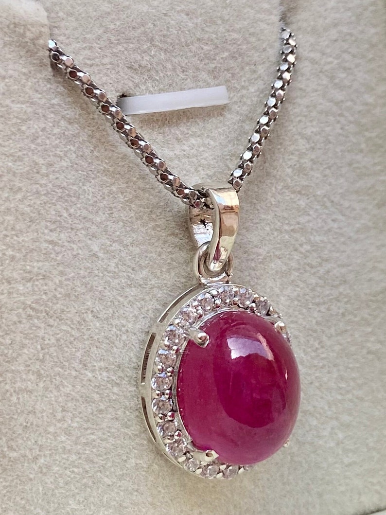 May include: A silver pendant necklace featuring a large, oval-shaped, deep red gemstone, surrounded by a halo of small, clear stones. The pendant hangs from a delicate, silver chain. The necklace is displayed against a light gray background.