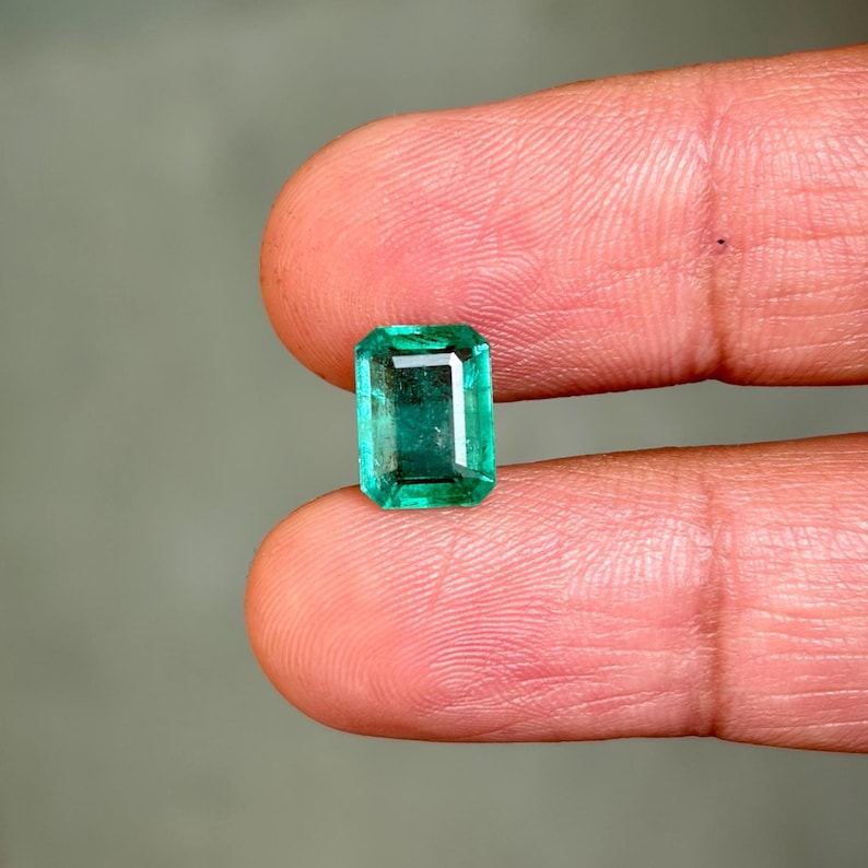 May include: A rectangular emerald gemstone with a vibrant green hue. The gem is held between two fingers, showcasing its clarity and cut. The emerald's facets reflect light, highlighting its brilliance. The background is a soft, neutral color.