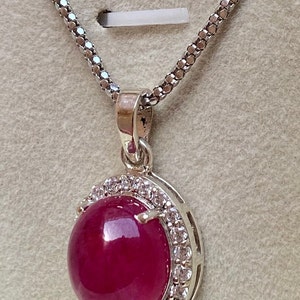 May include: A silver necklace with a round, deep red pendant. The pendant is surrounded by a halo of small, clear gemstones. The chain is a delicate, textured silver. The pendant is the focal point of the necklace.