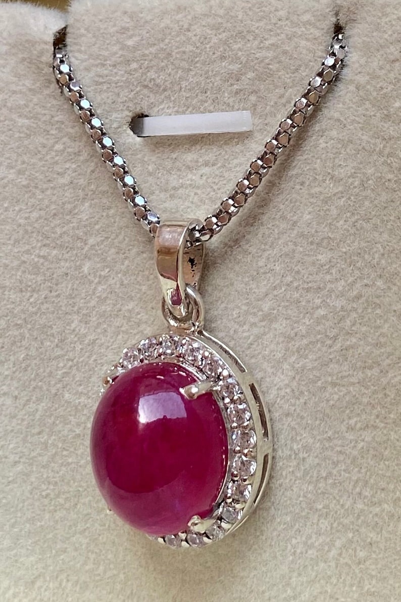 May include: A silver necklace with a round, deep red pendant. The pendant is surrounded by a halo of small, clear gemstones. The chain is a delicate, textured silver. The pendant is the focal point of the necklace.