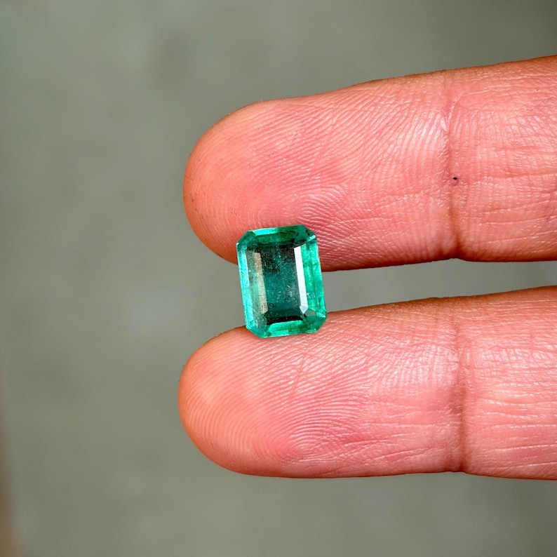 May include: A rectangular-cut emerald gemstone, showcasing a vibrant green hue. The gem is held between two fingers, highlighting its clarity and color. The emerald's facets reflect light, creating a sparkling effect. The background is a soft, neutral tone.