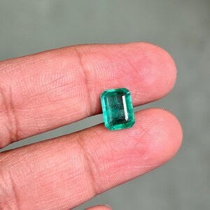 May include: A rectangular emerald gemstone with a vibrant green hue. The gem is held between two fingers, showcasing its polished facets and clarity. The emerald's shape is a classic rectangular cut, reflecting light and color.