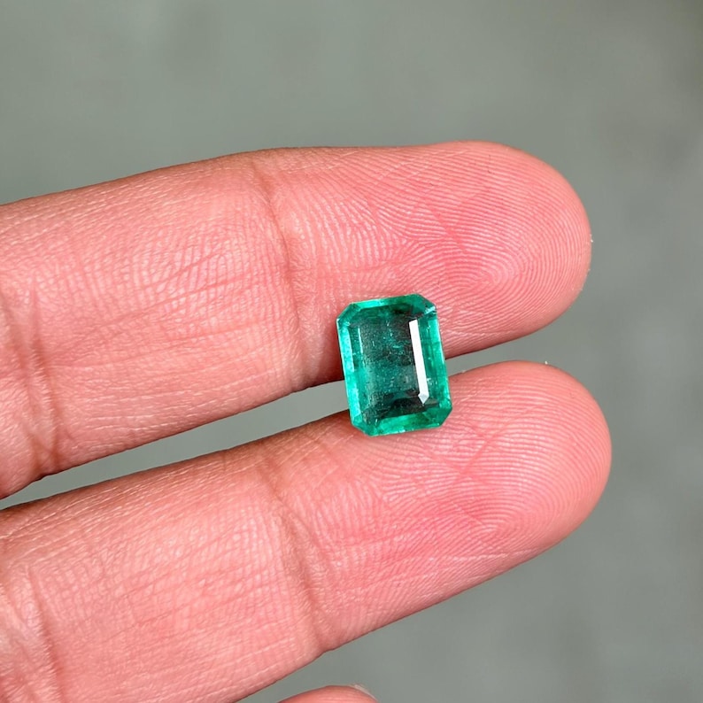 May include: A rectangular emerald gemstone with a vibrant green hue. The gem is held between two fingers, showcasing its polished facets and clarity. The emerald's shape is a classic rectangular cut, reflecting light and color.