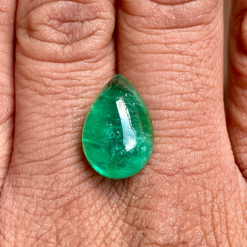 May include: A vibrant green emerald gemstone, cut in a teardrop shape, is displayed on a finger.