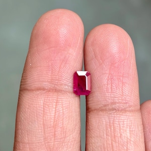 Puede incluir: Una gema roja vibrante de corte rectangular. La gema se sostiene entre dos dedos, mostrando sus facetas pulidas y su rico color. La forma de la piedra es un corte esmeralda clásico, que refleja la luz y muestra su brillo interno.